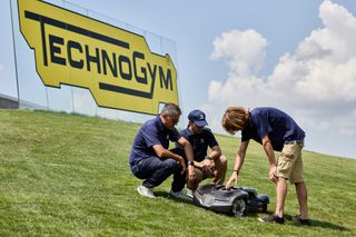 Tre persone che esaminano un robot tagliaerba su un pendio erboso con un'insegna Technogym sullo sfondo.