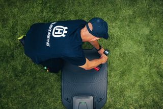 Osoba pracující na robotické sekačce na trávu na zahradě, oblečená v tričku značky Husqvarna.