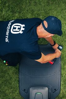 A person working on a robotic lawn mower in a garden, wearing a Husqvarna branded shirt.