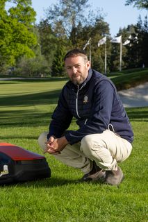 Osoba koja kleči pored robotske kosilice na golf terenu.