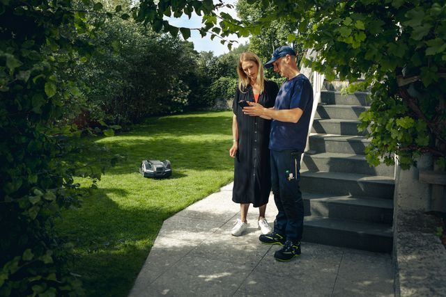 Two people standing on a patio, observing a robotic lawn mower operating in a garden.