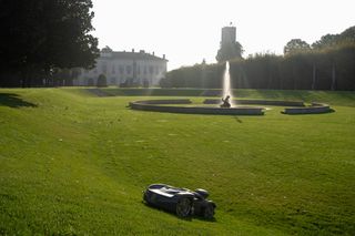 Robot tagliaerba al lavoro su un grande prato erboso con una fontana sullo sfondo.