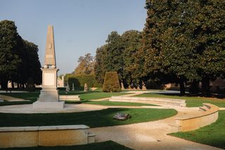 Robot tagliaerba che opera in un giardino formale con monumento a obelisco e siepi tagliate.