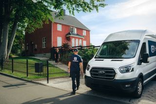 Eine Person in Arbeitskleidung, die auf ein rotes Haus zugeht, in dessen Nähe ein gebrandmarkter Lieferwagen geparkt ist.