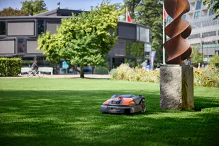 Robô corta-relva a funcionar num ambiente de parque com uma escultura e um edifício ao fundo.