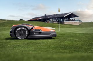 Robotska kosilica koja radi na golf terenu s klupskom kućom u pozadini.