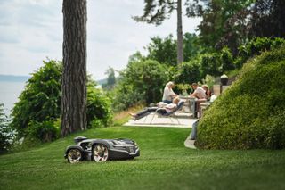 Robot tondeuse fonctionnant sur une pelouse herbeuse près d’un patio avec des gens qui se détendent.