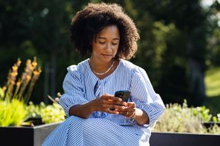 A person sitting outdoors on a sunny day, holding a smartphone.
