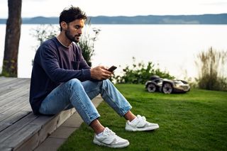 Uma pessoa sentada num deck de madeira, utilizando um smartphone, com um robô corta-relva a funcionar na relva nas proximidades.