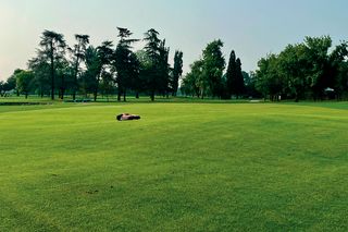 Robot tagliaerba al lavoro su un grande campo erboso ben curato con alberi sullo sfondo.