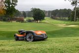 Robotic lawn mower operating on a golf course with trees and sand bunkers in the background.