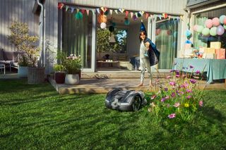 Een robotmaaier die in een tuin werkt met een persoon die zich in de buurt van een versierde patio bevindt.