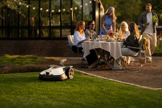 A group of people socializing in a garden with a robotic lawn mower operating nearby.