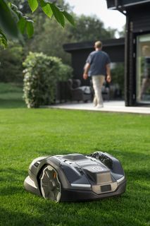 A robotic lawn mower operating on a well-maintained lawn with a person walking towards a modern house.