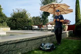En person som står på gräs och styr en robotgräsklippare med en fjärrkontroll i en trädgårdsmiljö.
