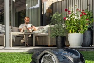 Mähroboter auf einem Rasen in der Nähe einer Terrasse mit einer Person im Hintergrund.