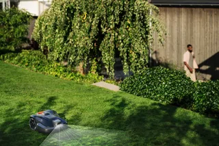 Robotic lawn mower operating in a garden with a blurred person in the background.