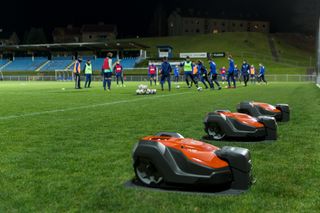 Deux robots tondeuses opérant sur un terrain de football la nuit, avec des joueurs s’entraînant en arrière-plan.