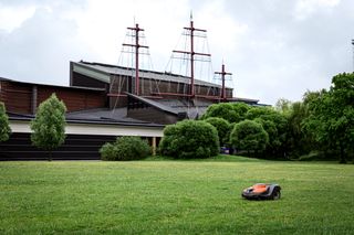 Robot tondeuse fonctionnant sur un champ herbeux près d’un bâtiment équipé de mâts de navire.