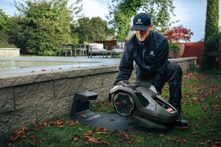 Osoba klečící vedle robotické sekačky na trávu poblíž bazénu, obklopená spadaným listím.