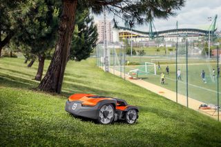 Robotic lawn mower operating on a grassy slope near a sports field.