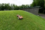 Robotic lawn mower operating on a grassy slope near a modern wooden building.