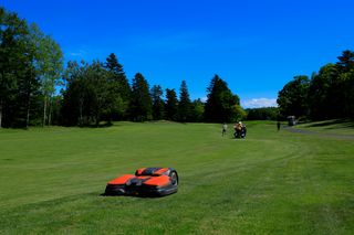 ゴルフコースのフェアウェイでのロボット芝刈機。人々とゴルフカートを背景に。