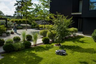 Een robotmaaier die werkt in een goed onderhouden tuin met een persoon die op de achtergrond de planten verzorgt.