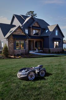 Robotic lawn mower operating on a grassy lawn in front of a large lakeside house.