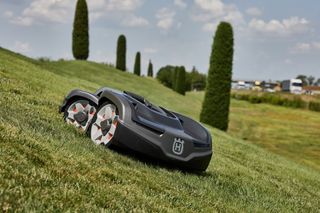 Robot tagliaerba che opera su un pendio erboso con alberi ad alto fusto sullo sfondo.