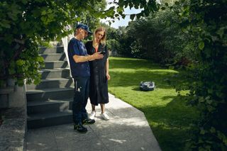 Two people standing on a patio, observing a robotic lawn mower operating in a garden.