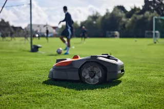 Robotic lawn mower operating on a sports field with people playing soccer in the background.