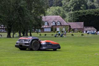 Robotic lawn mower operating on a large grassy field with houses in the background.
