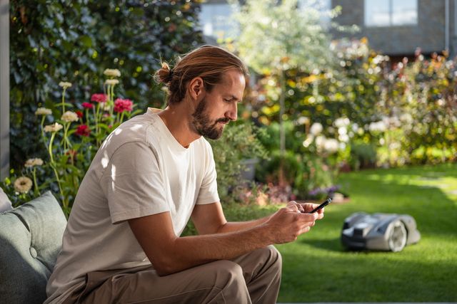 Eine Person, die mit einem Smartphone in einem Garten sitzt, während im Hintergrund ein Mähroboter läuft.