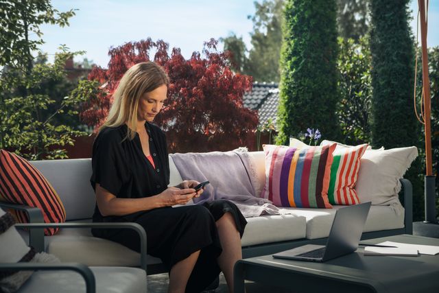 Eine Person, die mit einem Smartphone auf einer Terrasse sitzt, mit einem Laptop auf dem Tisch und bunten Kissen auf dem Sofa.