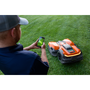 A person using a smartphone to control a robotic lawn mower on a grassy lawn.