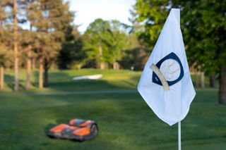 Un robot tondeuse circulant sur un parcours de golf à proximité d’un drapeau portant le logo d’un club de golf.