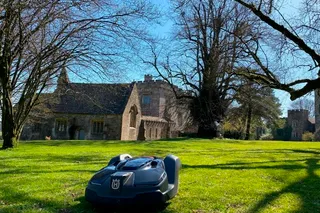 Robotic lawn mower operating on a sunny day in a garden with a historic building in the background.