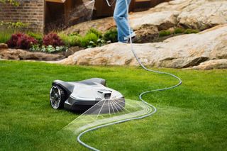 Un robot tondeuse fonctionnant sur une pelouse bien entretenue avec une personne en train d’arroser des plantes en arrière-plan.
