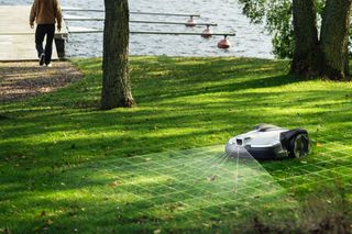 Un robot tondeuse opérant sur une zone herbeuse près d’un lac, avec une personne marchant vers un quai.