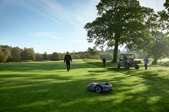 Kosiarka automatyczna pracująca na polu golfowym z ludźmi w tle.