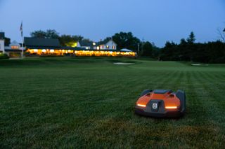 Robotic lawn mower operating on a golf course at dusk with a clubhouse in the background.