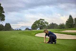 Une personne agenouillée sur un terrain de golf avec un robot tondeuse à proximité.