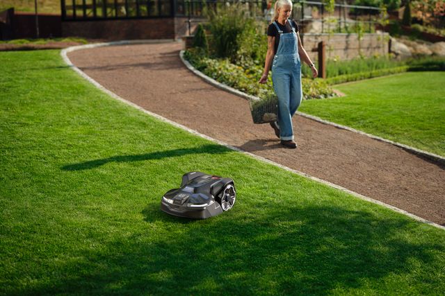 Robotic lawn mower cutting grass in a garden as a person walks on a path with a basket.