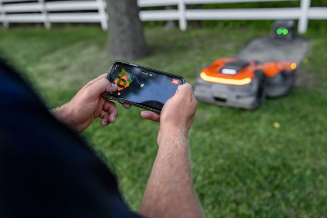 A person using a smartphone to control a robotic lawn mower in a garden.