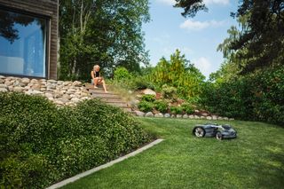 Un robot tondeuse fonctionnant sur une pente herbeuse près d’une maison moderne avec une personne assise sur les marches.