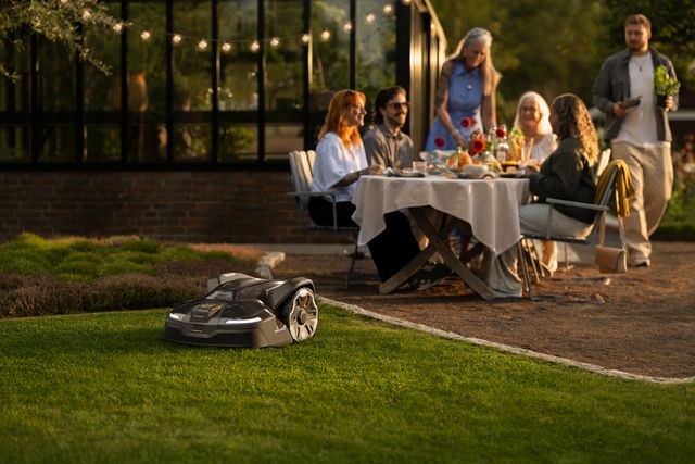 Robotic lawn mower operating on grass; group dining by a greenhouse in the background.