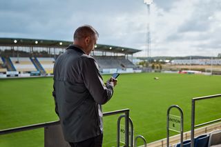 Een persoon die in een stadion staat, een smartphone vasthoudt en uitkijkt over het veld.
