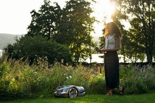 A person standing in a garden with a robotic lawn mower operating nearby.