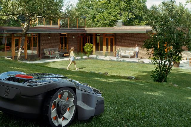 A robotic lawn mower operating on a grassy lawn in front of a modern house, with people in the background.
